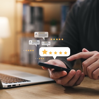 Eine-Person-sitzt-vor-seinem-Laptop-tippt-in-sein-smartphone-eine-sehr-gute-bewertung-ein-im-hintergrund-tauchen-andere-bewertungen-aus-seinen-Smartphone-auf-daneben-steht-ein-Laptop-auf-dem-Tisch